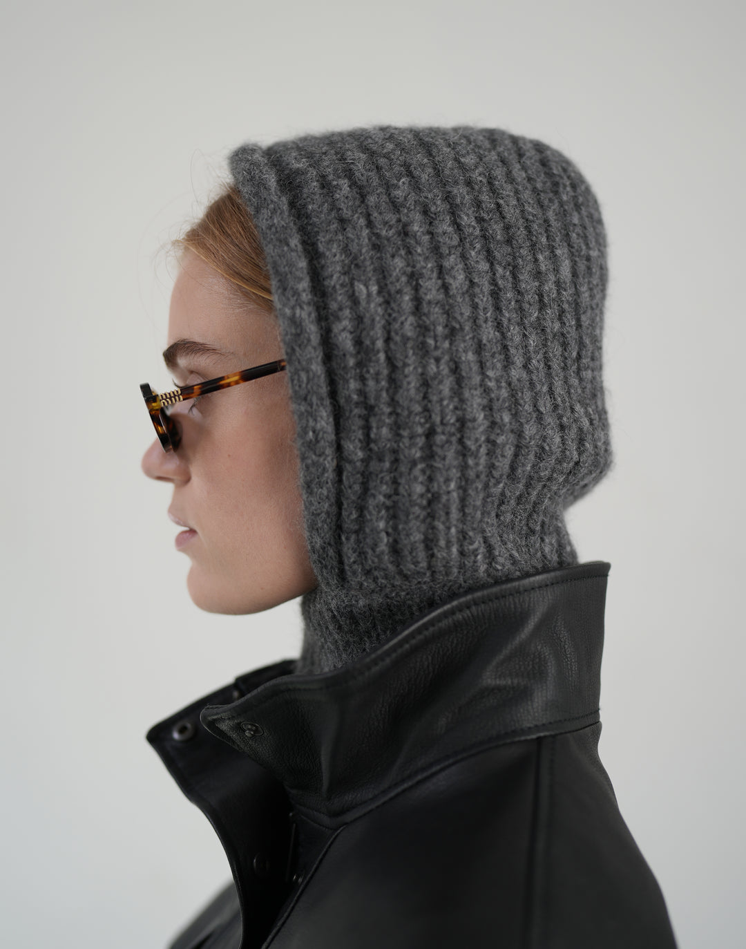 A side-profile view of a person wearing a dark gray ribbed knit hooded balaclava with a high neck, styled with tortoiseshell glasses and a black leather jacket against a light gray background. #color_grey