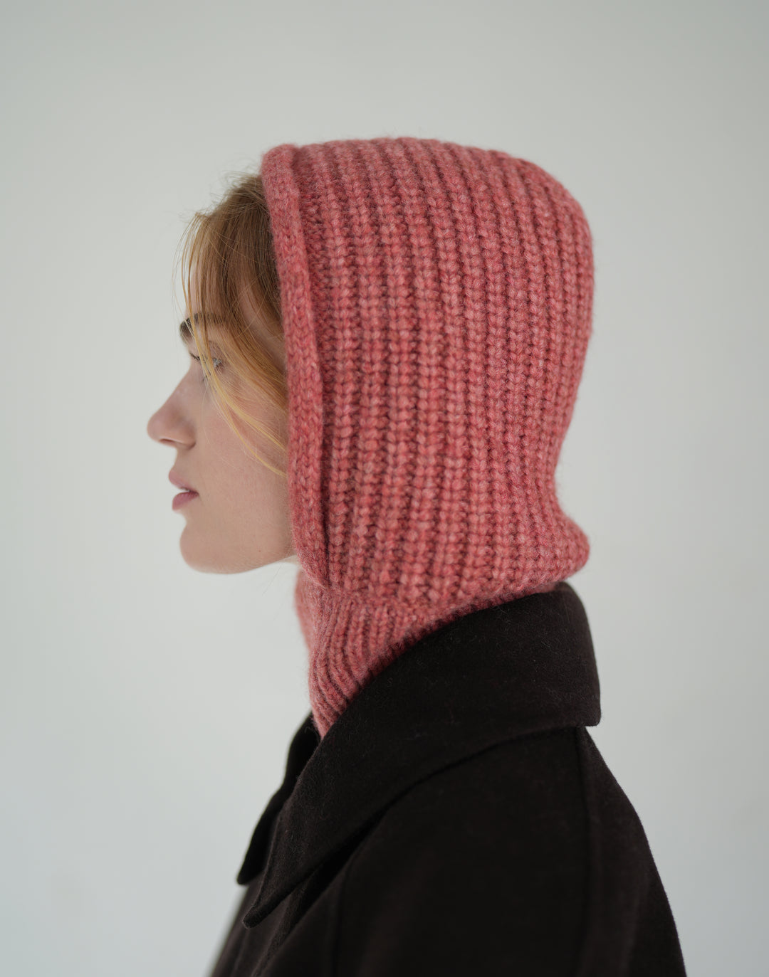 A person shown in side profile wearing a chunky knit hood in dusty rose pink over a dark coat, against a plain light gray background. #color_dusty-pink