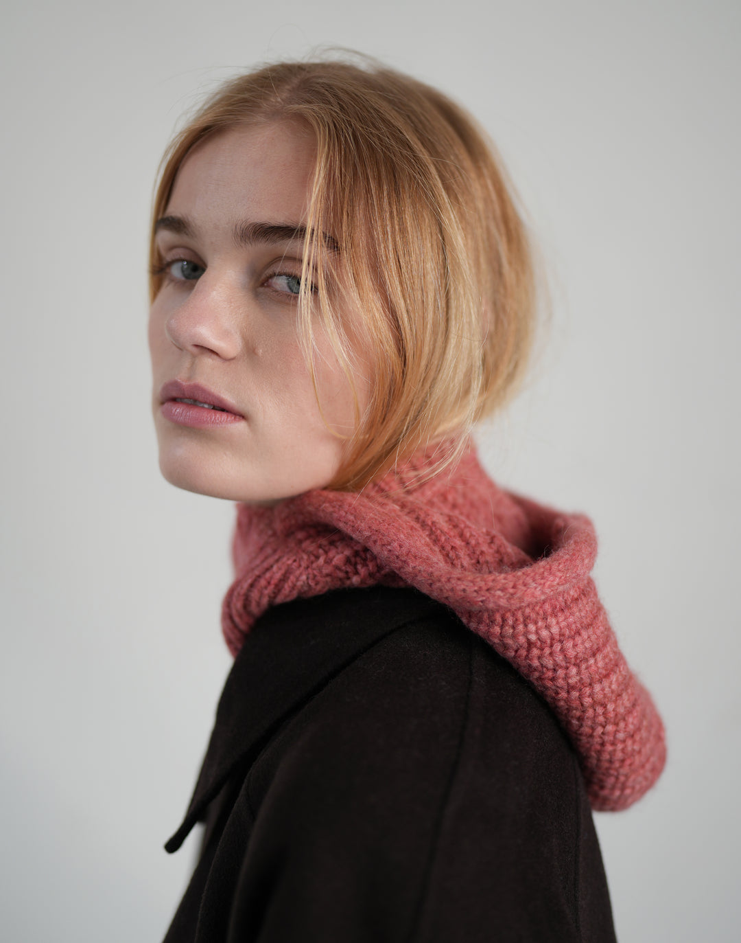 A woman with blonde hair wearing a chunky knit cowl in a dusty rose pink color, styled over a dark brown coat against a plain light gray background. #color_dusty-pink