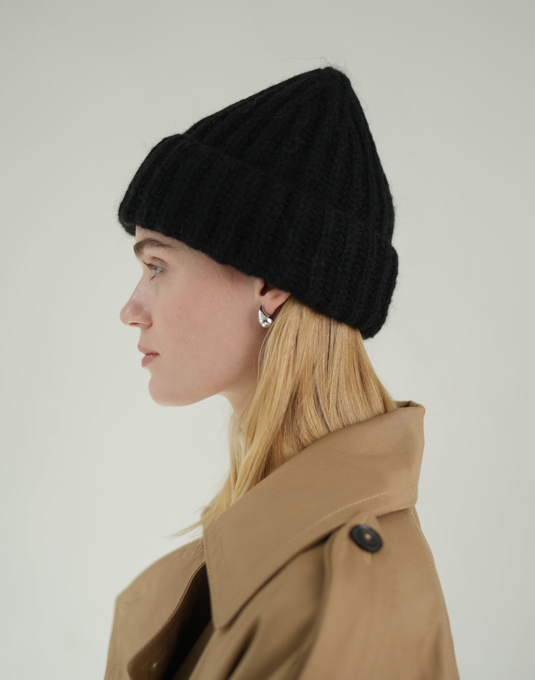 A woman in profile wearing a black ribbed knit beanie and a tan trench coat against a plain light gray background. #color_black