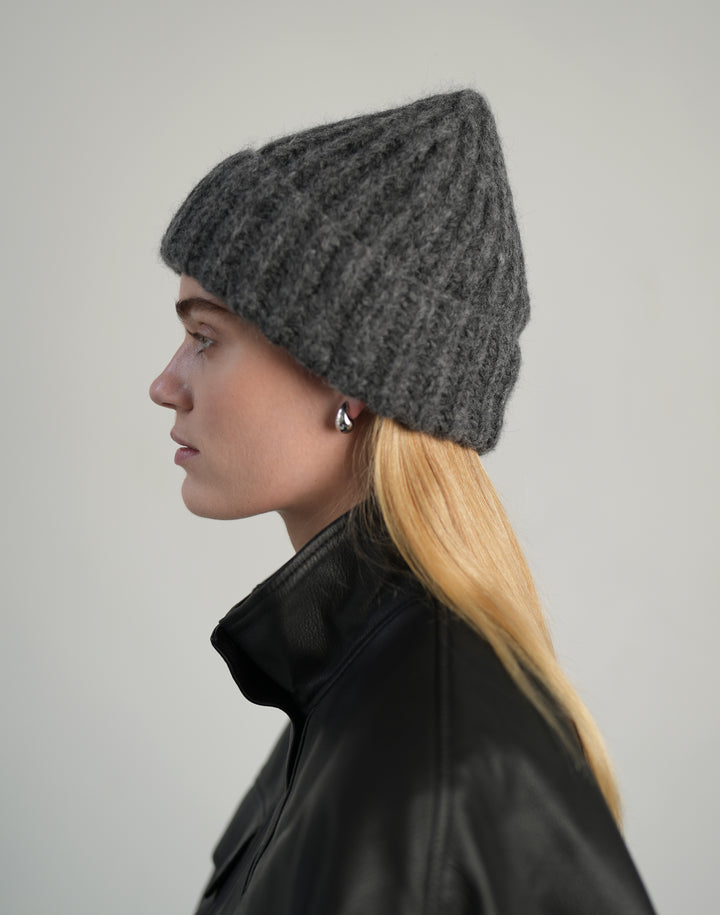 A woman in profile wearing a dark gray ribbed knit beanie and a black leather jacket, photographed against a plain light gray studio background. #color_grey