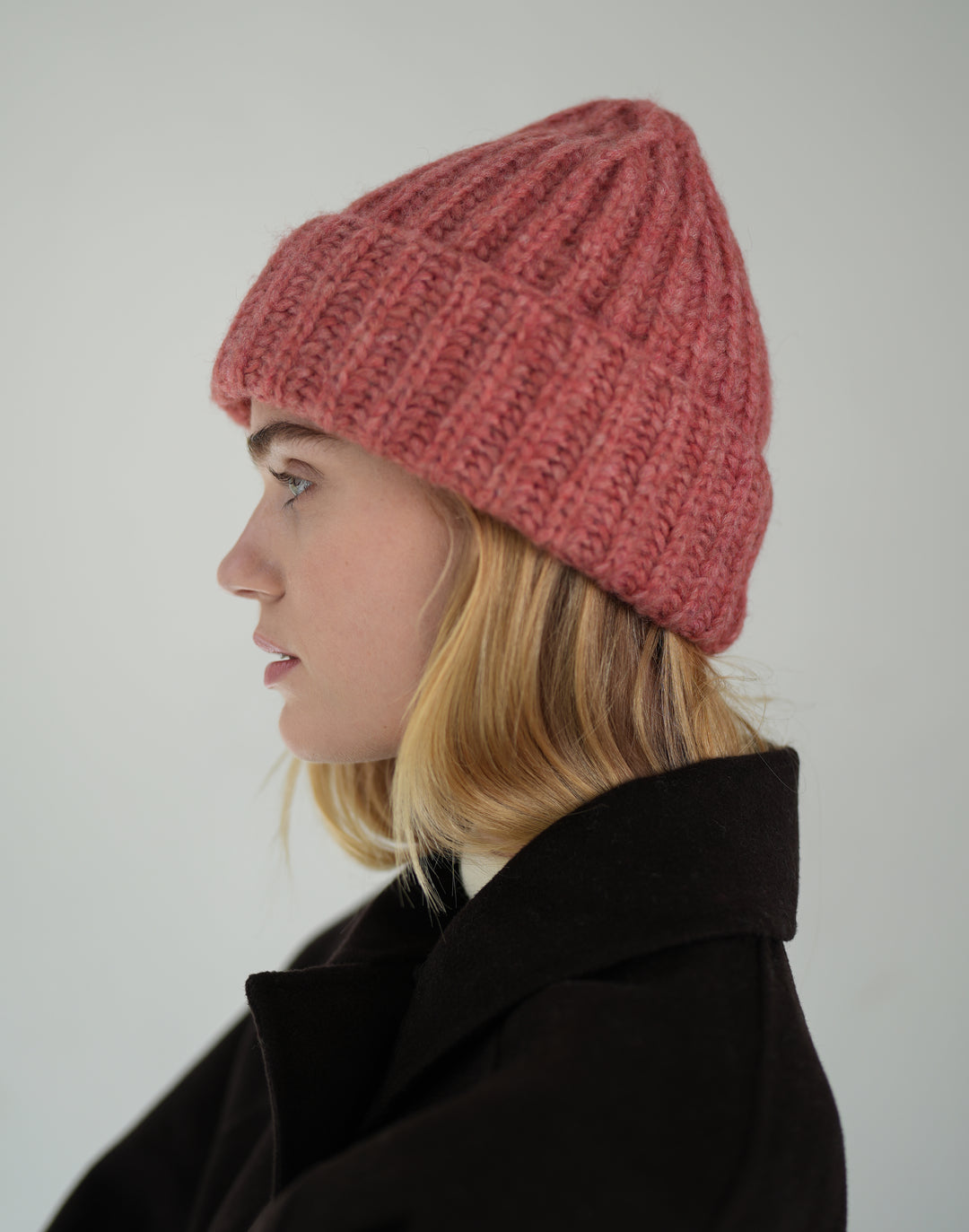 A woman in profile wearing a chunky knit beanie in a dusty rose pink color, paired with a dark coat, against a plain light gray background. #color_dusty-pink