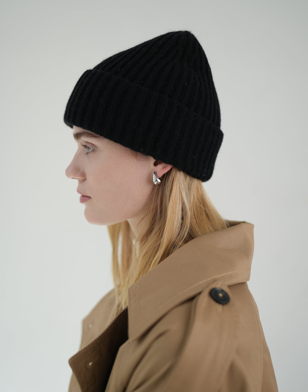 A woman in profile wearing a black ribbed knit beanie and a tan trench coat, photographed against a plain light gray studio background.#color_black