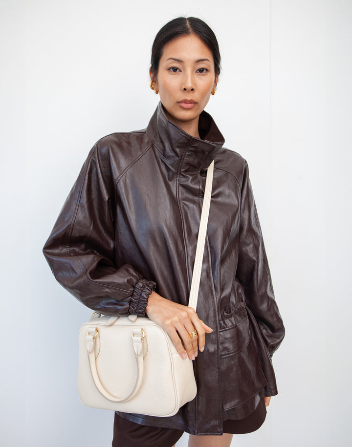 a model wears a hyer goods la corsa off-white leather bowling bag bone with gold hardware as a crossbody bag #color_bone
