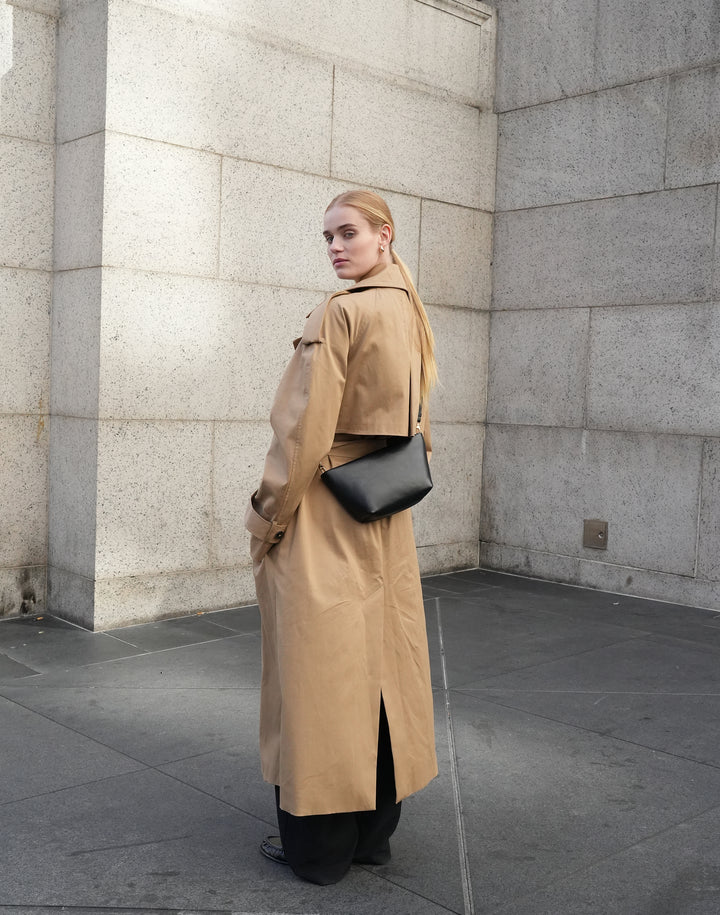 A woman wearing a long tan trench coat and black pants stands in a stone corridor, carrying a black shoulder bag. #color_black-silver