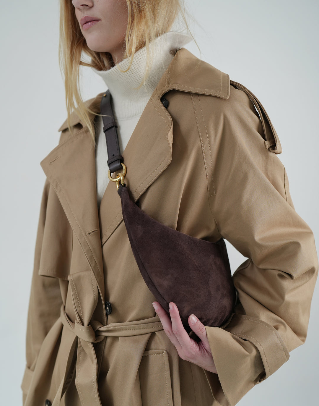 a cropped photo of a womans torso- she wears a cream turtleneck, khaki belted trench and a dark brown suede crossbody crescent bag with leather strap #color_chocolate-suede