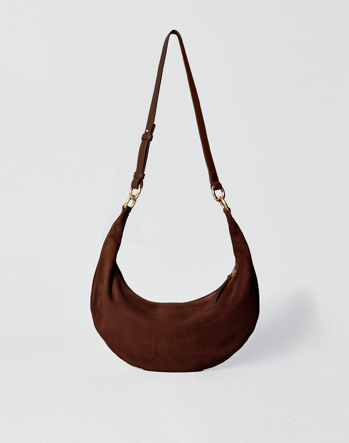 A dark brown HYER GOODS Piccola Ring Bag crescent-shaped shoulder bag made of suede, featuring a single adjustable shoulder strap with gold-tone hardware, set against a light gray background #color_chocolate-suede