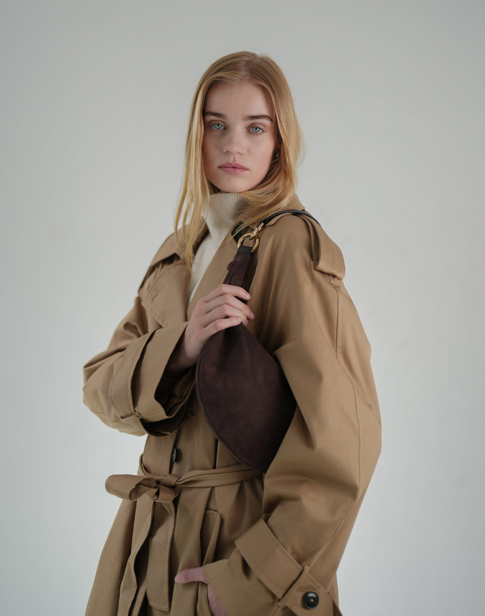 a woman wears a cream sweater turtleneck and a belted, oversized khaki trench coat with a brown suede crescent shoulder bag #color_chocolate-suede