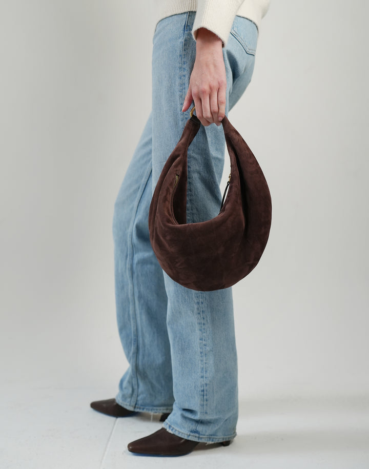 a cropped photo of a womans light blue jeans, brown pointy leather boots and chocolate suede crescent bag hanging from her fingertips #color_chocolate-suede