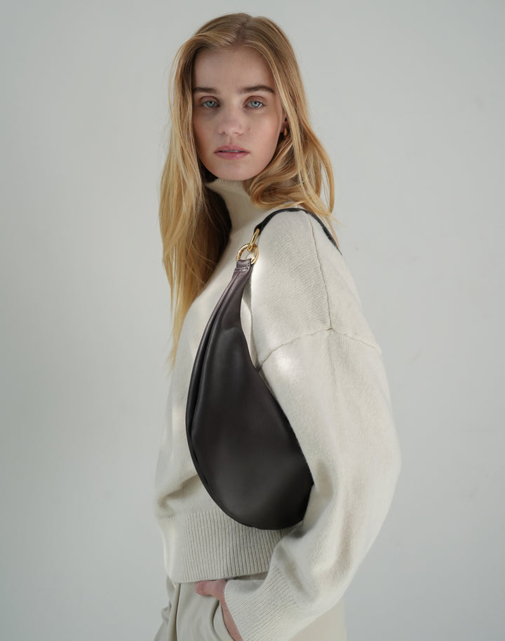 profile shot of a woman wearing a cream oversized turtlneck sweater and a brown leather crescent shoulder bag #color_chocolate
