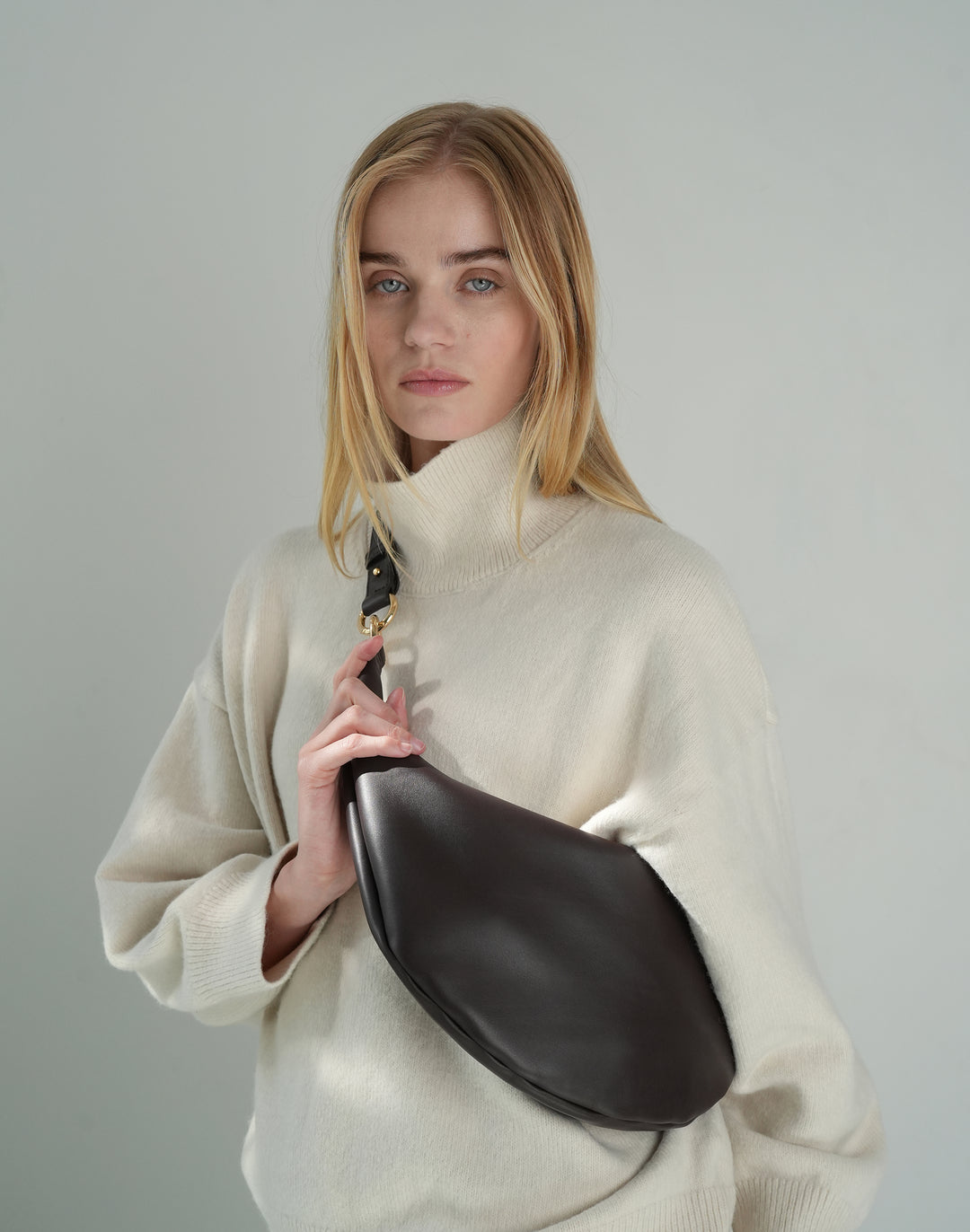 a woman wears an oversized cream turtleneck sweater with a dark brown leather crossbody crescent bag slung on top. #color_chocolate