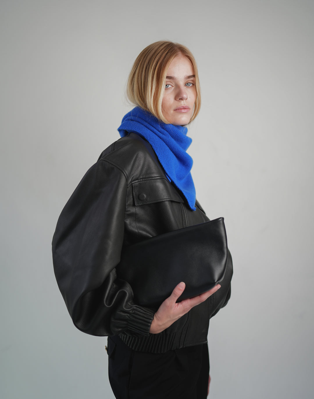 A model wearing a black leather jacket, black pants, and a bright blue knit scarf, holding a black leather clutch bag against a solid gray studio background.#color_cobalt