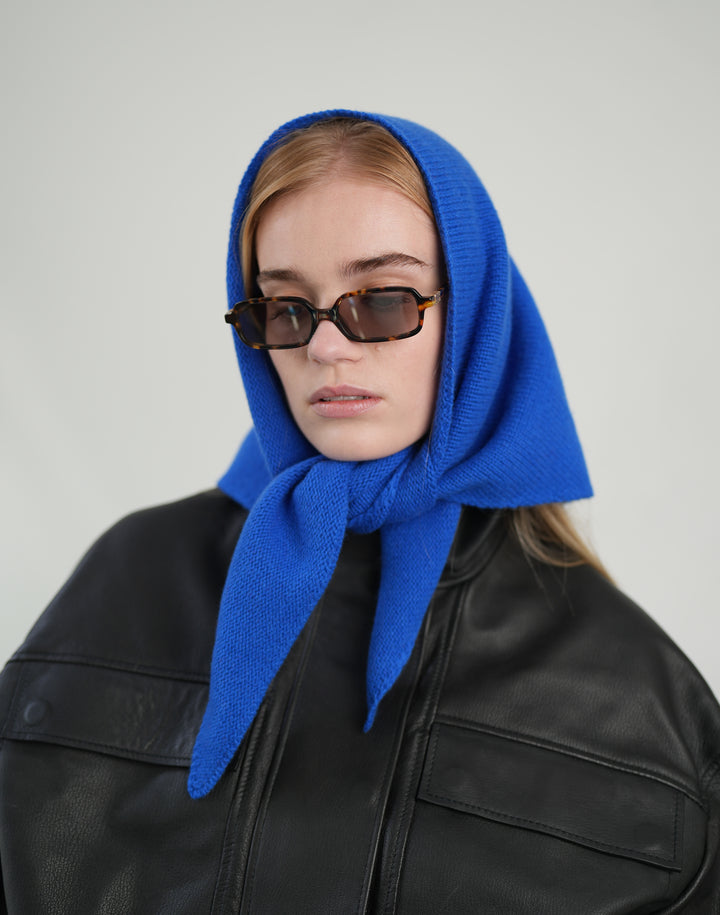 A model wearing a bright royal blue knitted headscarf tied under the chin, styled with dark sunglasses and a black leather jacket against a plain light gray background.#color_cobalt