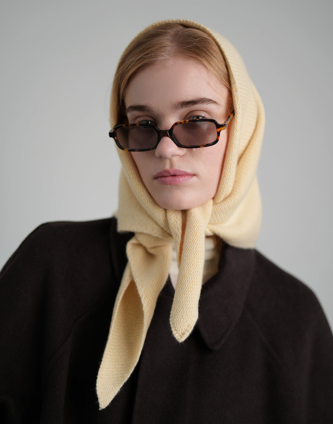 A model wearing a cream-colored knitted headscarf tied under the chin, dark tortoiseshell sunglasses, and a dark brown coat against a solid gray background. #color_butter-yellow