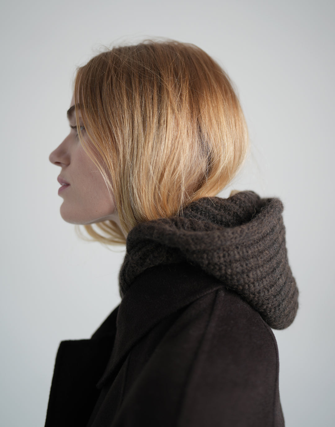 A side-profile portrait of a woman with blonde hair wearing a dark brown chunky knit cowl scarf and a dark coat against a light gray background. #color_brown