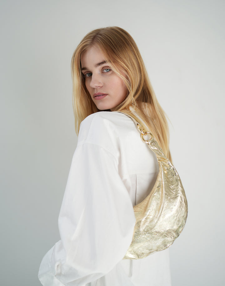 a blond-haired woman wearing a white oversized buttondown and a gold leather crescent shoulder bag #color_gold