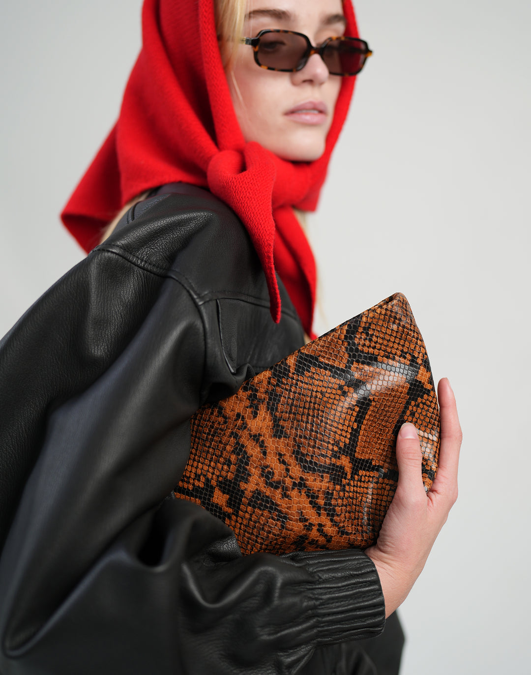 a cropped photo of a model in a red head scarf, oversized leather jacket, clutching a tan python clutch under her arm  #color_tan-python
